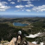 woman sitting alone on mountain summit overlooking alpine lake, solo travel photo taken with phone tripod