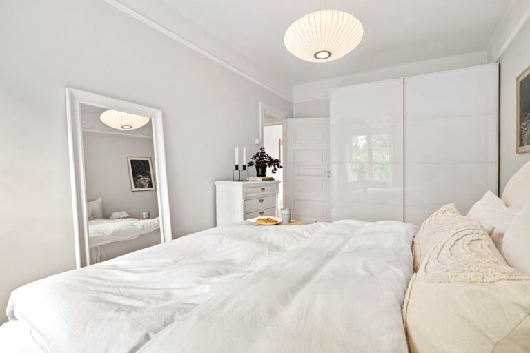 Bright minimalist bedroom with full-length floor mirror, dresser with plant and candles, white linen bedding