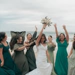 Women in sage green and teal beach wedding guest outfits celebrating together on a sunny coastal beach