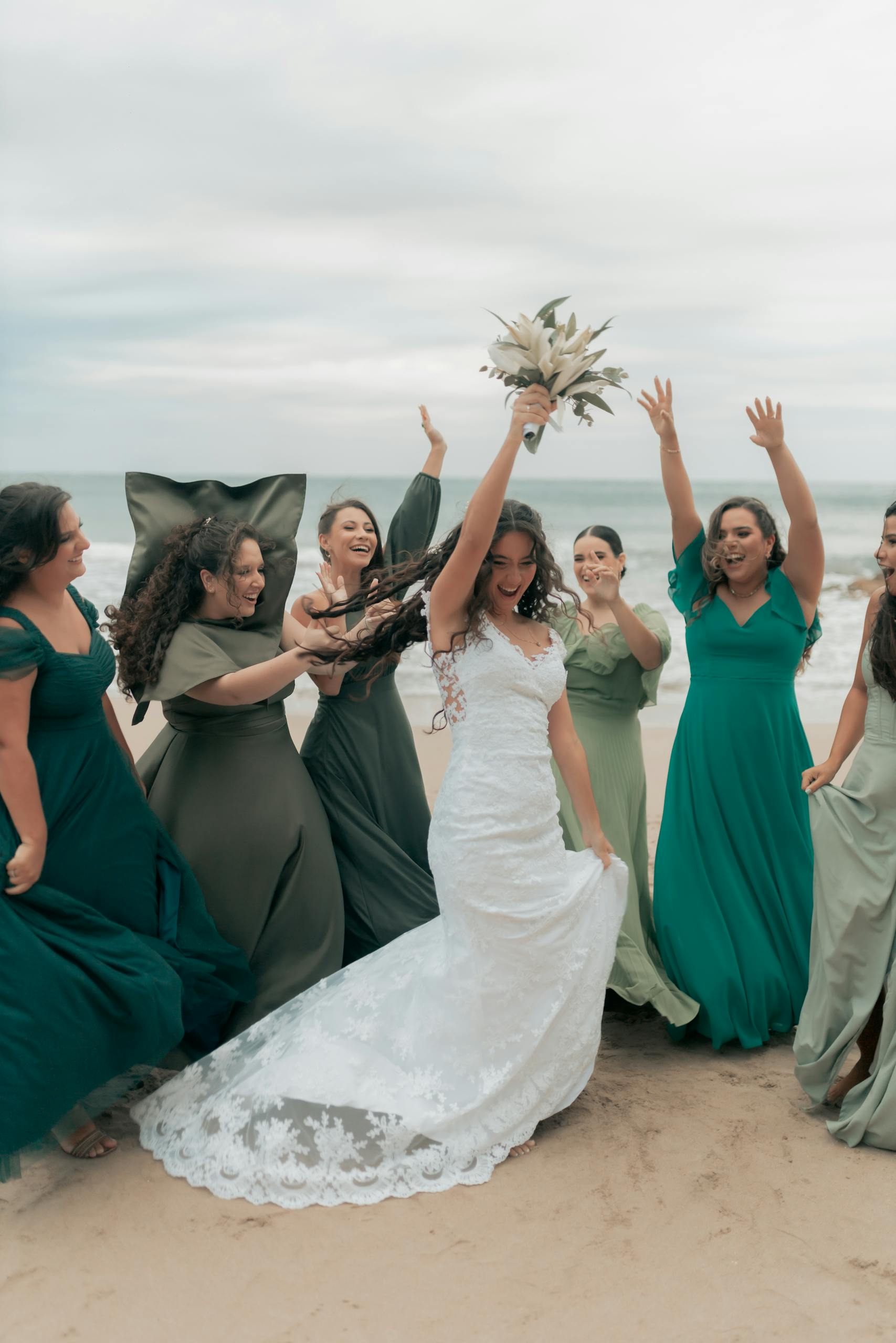 Women in sage green and teal beach wedding guest outfits celebrating together on a sunny coastal beach